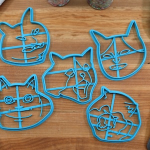 May include: Five blue plastic cookie cutters shaped like different cat faces. The cutters are on a wooden surface.