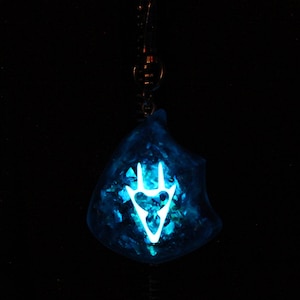 May include: A blue resin pendant with a white glow-in-the-dark symbol. The symbol is a stylized hand with three fingers.