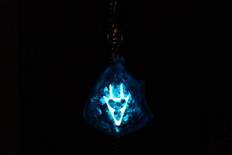 May include: A blue resin pendant with a white glow-in-the-dark symbol. The symbol is a stylized hand with three fingers.