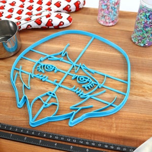 May include: A blue plastic cookie cutter shaped like a cartoon face with large eyes and a wide mouth. The cutter is on a wooden surface with a ruler, measuring cups, and sprinkles.