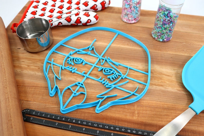 May include: A blue plastic cookie cutter shaped like a cartoon face with large eyes and a wide mouth. The cutter is on a wooden surface with a ruler, measuring cups, and sprinkles.