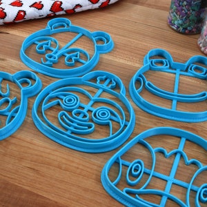 May include: Five blue plastic cookie cutters shaped like animal faces. The cutters are on a wooden surface. The cutters are for making cookies in the shapes of a bear, a panda, a frog, a sloth, and a turtle.