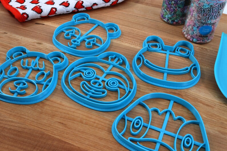 May include: Five blue plastic cookie cutters shaped like animal faces. The cutters are on a wooden surface. The cutters are for making cookies in the shapes of a bear, a panda, a frog, a sloth, and a turtle.