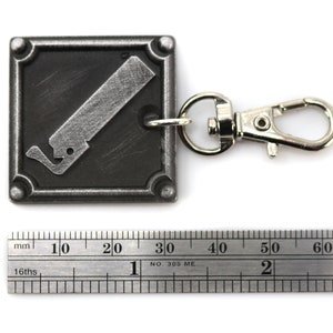 May include: A silver metal keychain with a black square charm featuring a stylized image of a saw blade. The charm has a silver border and four raised rivets. The keychain is attached to a silver metal split ring.