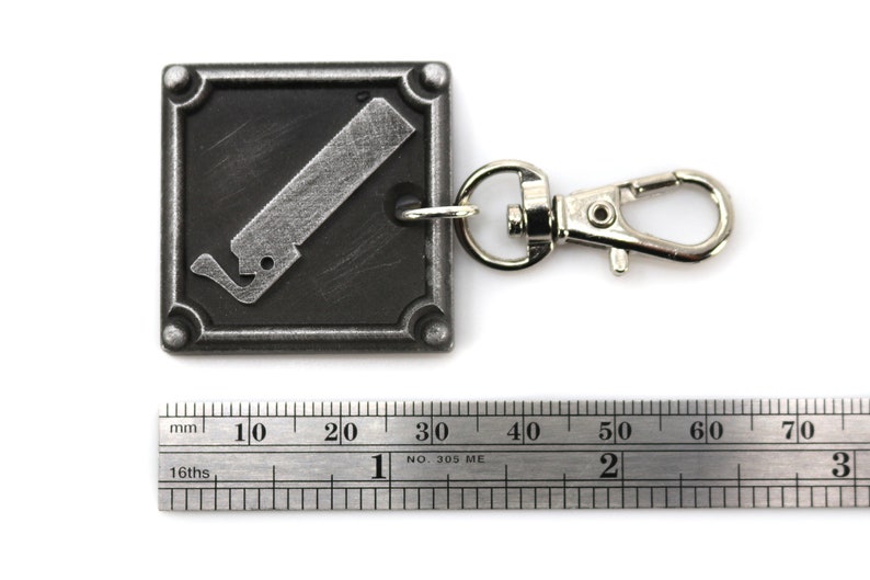 May include: A silver metal keychain with a black square charm featuring a stylized image of a saw blade. The charm has a silver border and four raised rivets. The keychain is attached to a silver metal split ring.