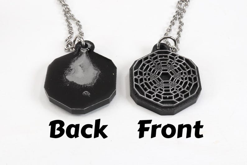 May include: A silver chain necklace with a black octagonal pendant. The front of the pendant features a silver geometric design. The back of the pendant is plain.