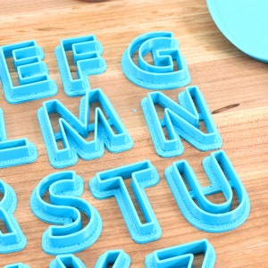May include: A set of blue plastic alphabet cookie cutters on a wooden surface. The cutters are arranged in alphabetical order, starting with the letter D and ending with the letter Z.