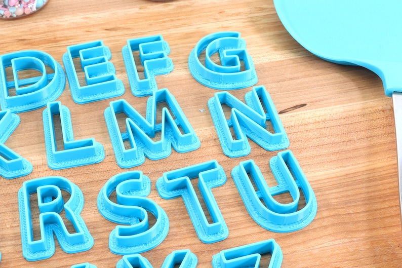 May include: A set of blue plastic alphabet cookie cutters on a wooden surface. The cutters are arranged in alphabetical order, starting with the letter D and ending with the letter Z.
