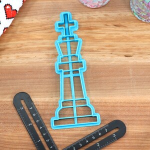 May include: A blue plastic cookie cutter shaped like a chess king piece. The cutter is on a wooden surface.