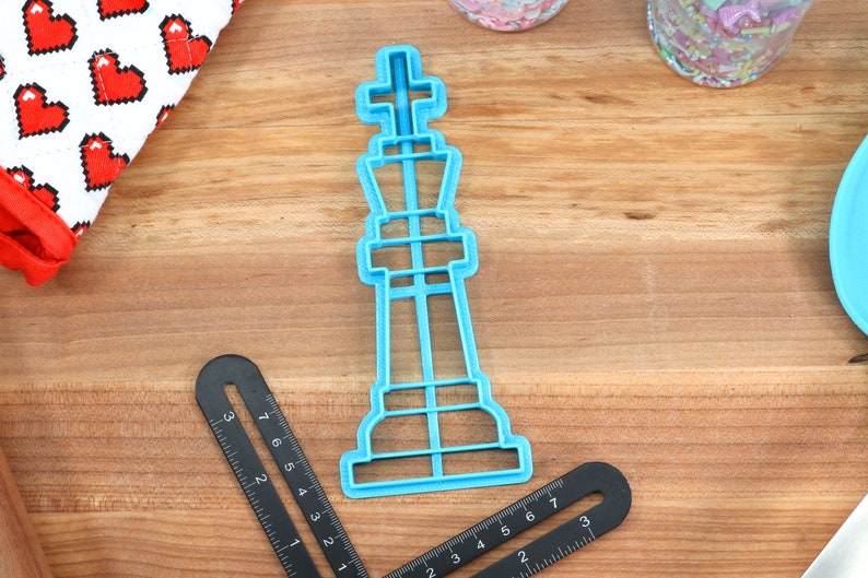 May include: A blue plastic cookie cutter shaped like a chess king piece. The cutter is on a wooden surface.