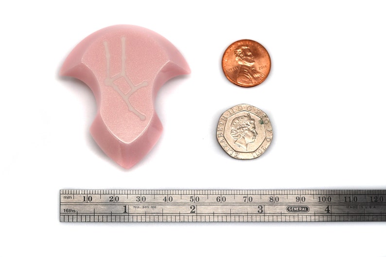 May include: A pink, irregularly shaped, ceramic pendant with a white design of the Gemini constellation. The pendant is shown next to a ruler, a US penny, and a British coin for size comparison.