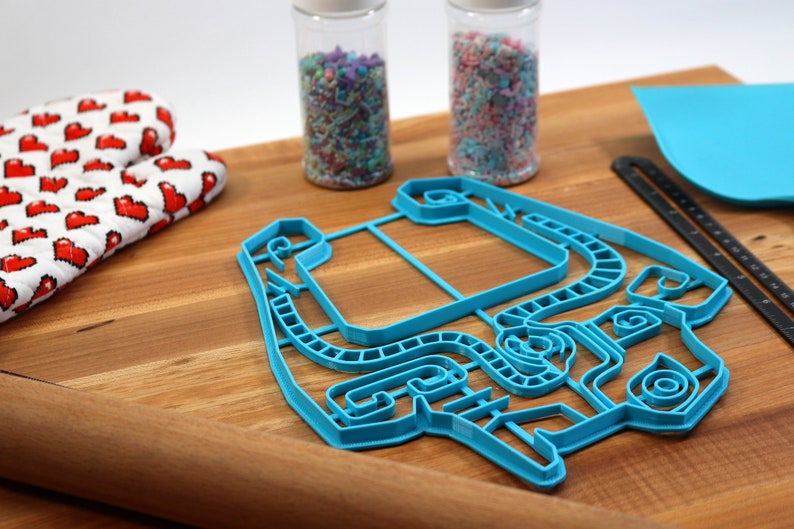 Peut inclure: Un emporte-pi&egrave;ce en plastique bleu en forme de personne avec un design d&eacute;taill&eacute;. L'emporte-pi&egrave;ce est sur une surface en bois avec un rouleau &agrave; p&acirc;tisserie, des vermicelles dans des bocaux et un tapis de d&eacute;coupe bleu.