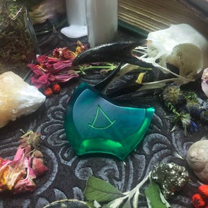 May include: A green, triangular-shaped pendant with a white symbol in the center, surrounded by dried flowers, a crystal, and a bird skull.