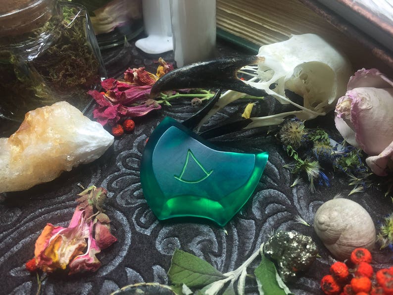 May include: A green, triangular-shaped pendant with a white symbol in the center, surrounded by dried flowers, a crystal, and a bird skull.