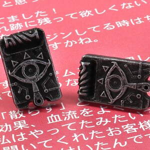 May include: Two silver-colored metal earrings shaped like a video game item with a silver eye and a triangle design. The earrings are on a red background.