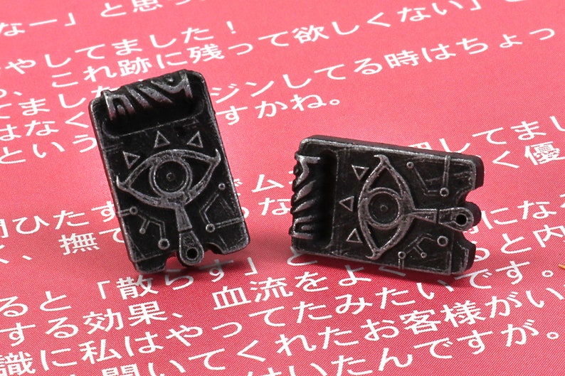 May include: Two silver-colored metal earrings shaped like a video game item with a silver eye and a triangle design. The earrings are on a red background.