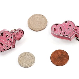 May include: Two pink heart-shaped pins with the text "DORK DORK CLUB" on them. The pins are on a white background.