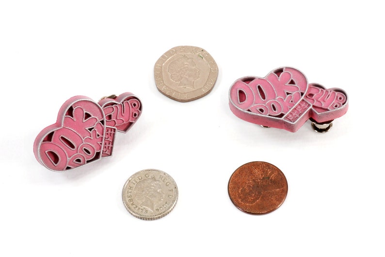 May include: Two pink heart-shaped pins with the text "DORK DORK CLUB" on them. The pins are on a white background.