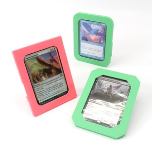 Op de afbeelding: Drie verschillende gekleurde kunststof kaarthouders met een kaart in elke houder. De houders zijn roze, groen en lichtgroen. De kaarten zijn allemaal Magic: The Gathering-kaarten.
