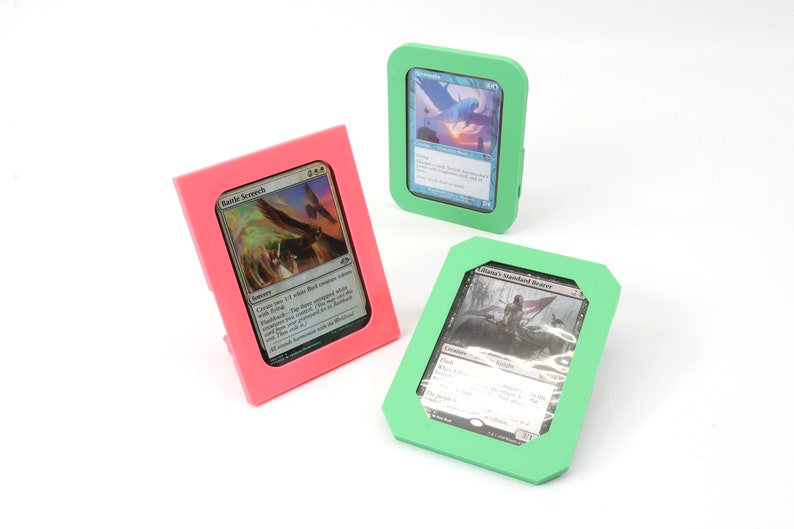Op de afbeelding: Drie verschillende gekleurde kunststof kaarthouders met een kaart in elke houder. De houders zijn roze, groen en lichtgroen. De kaarten zijn allemaal Magic: The Gathering-kaarten.