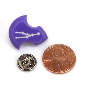 Op de afbeelding: Paarse emaille pin met een wit sterrenbeeld ontwerp. De pin is schildvormig en heeft een zilveren pin achterkant. Een cent is inbegrepen voor groottevergelijking.