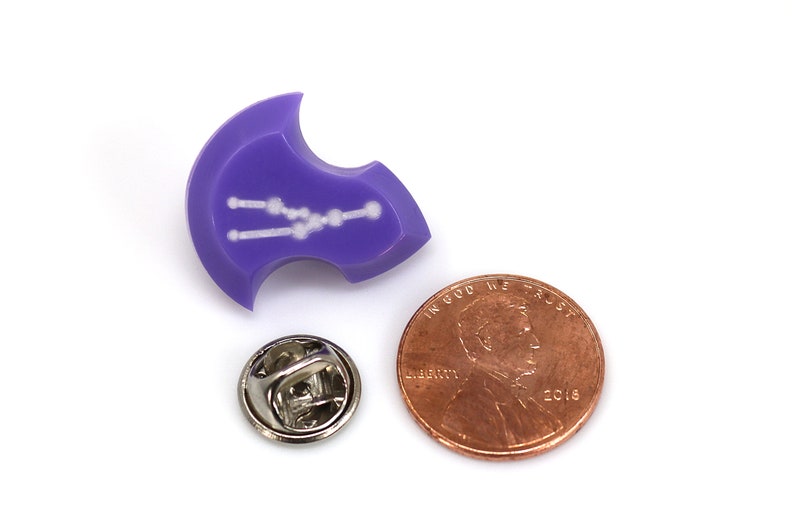 Op de afbeelding: Paarse emaille pin met een wit sterrenbeeld ontwerp. De pin is schildvormig en heeft een zilveren pin achterkant. Een cent is inbegrepen voor groottevergelijking.