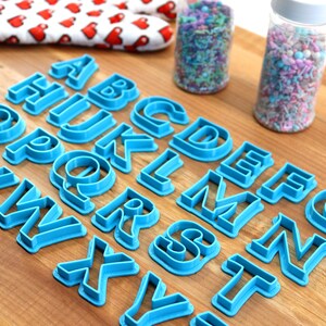 May include: A set of blue plastic alphabet cookie cutters on a wooden surface. The cutters are arranged in alphabetical order.