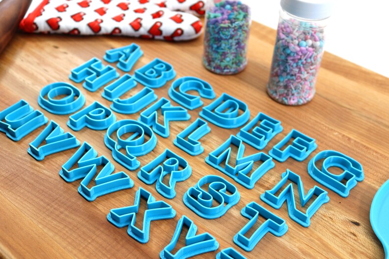 May include: A set of blue plastic alphabet cookie cutters on a wooden surface. The cutters are arranged in alphabetical order.