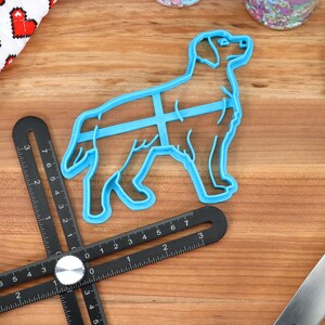 May include: A blue plastic dog-shaped cookie cutter on a wooden surface. The cutter is next to a black ruler with white markings.