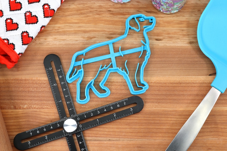 May include: A blue plastic dog-shaped cookie cutter on a wooden surface. The cutter is next to a black ruler with white markings.