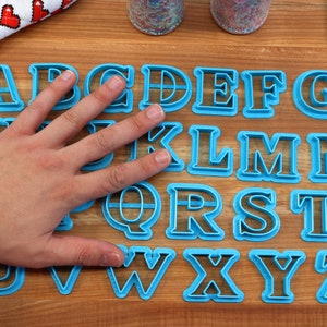 May include: A set of 26 blue plastic alphabet cookie cutters on a wooden surface. The cutters are arranged in alphabetical order.