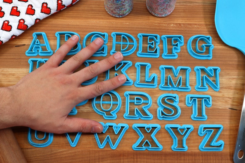 May include: A set of 26 blue plastic alphabet cookie cutters on a wooden surface. The cutters are arranged in alphabetical order.