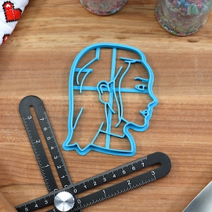 Op de afbeelding: Een blauwe plastic koekjesvormer in de vorm van een vrouwenhoofd met lang haar. De vormer ligt op een houten oppervlak.