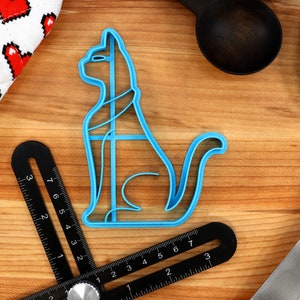 May include: A blue plastic cat-shaped cookie cutter on a wooden surface. The cutter has a detailed design and is perfect for making cat-shaped cookies.