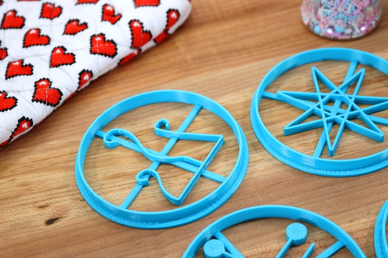 May include: Four blue plastic cookie cutters on a wooden surface. The cutters are shaped like a star, a bell, a geometric design, and a circle with a line through it.