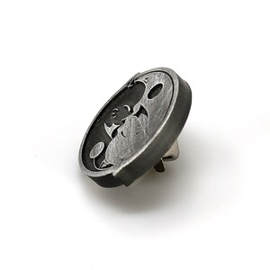 May include: A silver-toned metal pin with a black and white design of a cartoon character. The character is a creature with large eyes and a wide mouth.