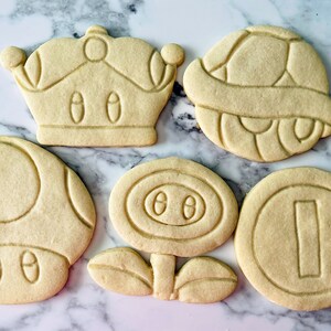 May include: Five sugar cookies decorated with icing in the shapes of a mushroom, a flower, a coin, a crown, and a turtle shell.