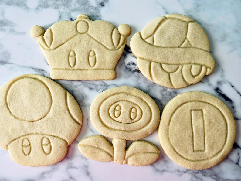 May include: Five sugar cookies decorated with icing in the shapes of a mushroom, a flower, a coin, a crown, and a turtle shell.