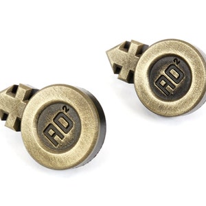 May include: Two gold-colored metal buttons with a black background. Each button features a stylized gender symbol and the text "AD2".