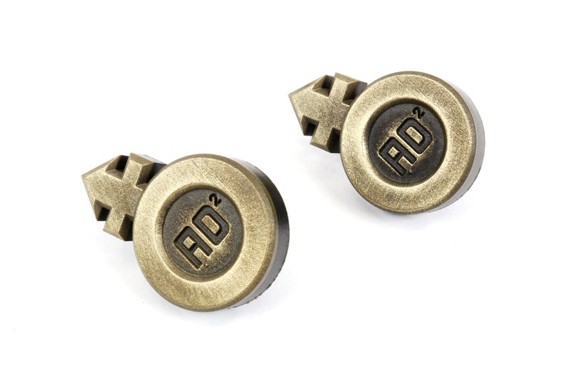 May include: Two gold-colored metal buttons with a black background. Each button features a stylized gender symbol and the text "AD2".