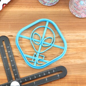 May include: A blue plastic cookie cutter shaped like an alien head with a square frame. The cookie cutter is on a wooden surface.