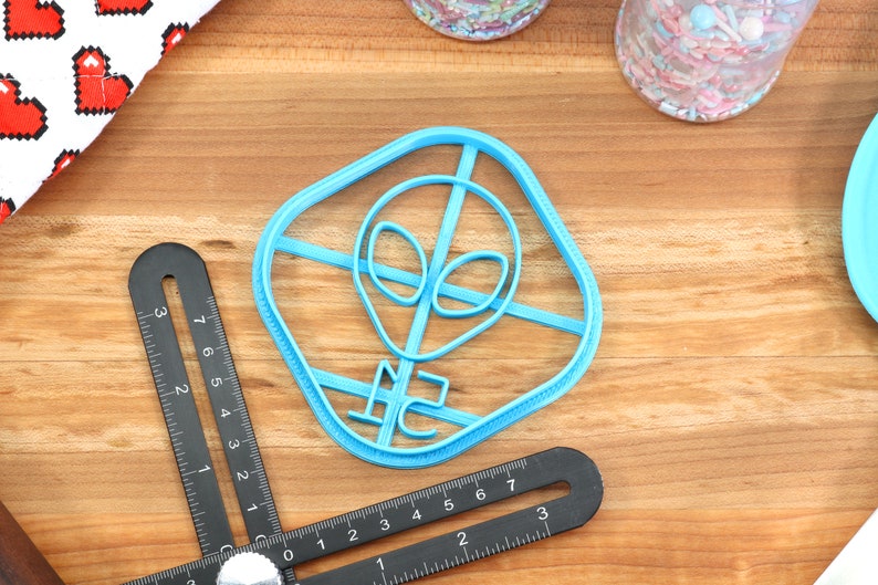 May include: A blue plastic cookie cutter shaped like an alien head with a square frame. The cookie cutter is on a wooden surface.
