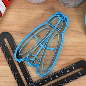 May include: A blue plastic cookie cutter shaped like a dragonfly. The cutter has a detailed design with intricate wings and a body.
