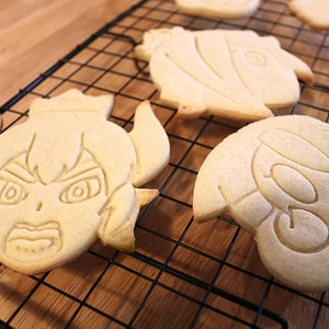 May include: Four decorated sugar cookies on a black wire cooling rack. The cookies are shaped like cartoon characters with detailed facial features.