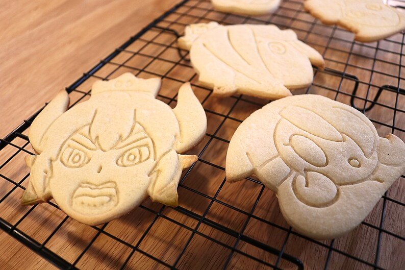 May include: Four decorated sugar cookies on a black wire cooling rack. The cookies are shaped like cartoon characters with detailed facial features.
