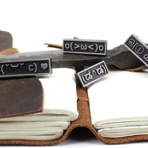 May include: Four silver-toned metal bookmarks with different symbols and text. The bookmarks are on a brown leather journal and a metal surface. The text on the bookmarks includes: "( ˘ &sup3;˘)&hearts;", "o(>&omega;<)o", "( ˘&omega;˘ )", and "( ಡ ﹏ ಡ)".