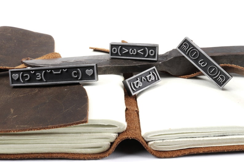 May include: Four silver-toned metal bookmarks with different symbols and text. The bookmarks are on a brown leather journal and a metal surface. The text on the bookmarks includes: "( ˘ &sup3;˘)&hearts;", "o(>&omega;<)o", "( ˘&omega;˘ )", and "( ಡ ﹏ ಡ)".