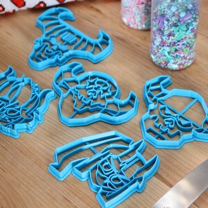 May include: Five blue plastic cookie cutters shaped like different fantasy creatures. The cutters are on a wooden surface with a jar of sprinkles in the background.