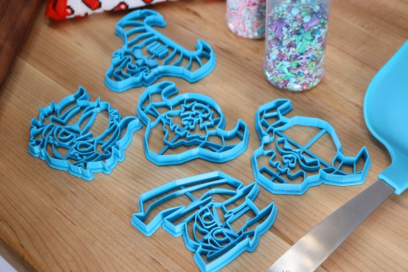 May include: Five blue plastic cookie cutters shaped like different fantasy creatures. The cutters are on a wooden surface with a jar of sprinkles in the background.