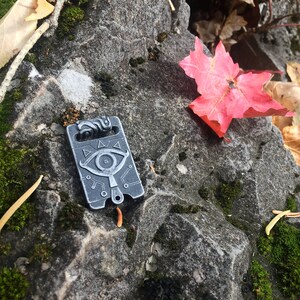 May include: A silver metal charm with a stylised eye design and geometric patterns. The charm is on a rocky surface with a red maple leaf.